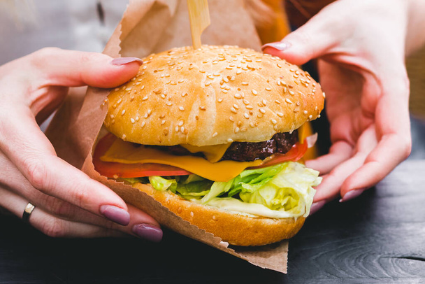 Ženské ruce drží čerstvý lahodný burger s hranolky na černém dřevěném stole - Fotografie, Obrázek