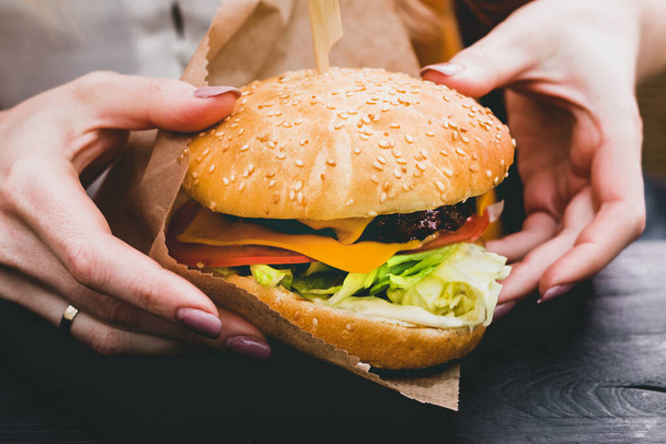 Руки женщины держат свежий вкусный бургер с картошкой фри на черном деревянном столе - Фото, изображение