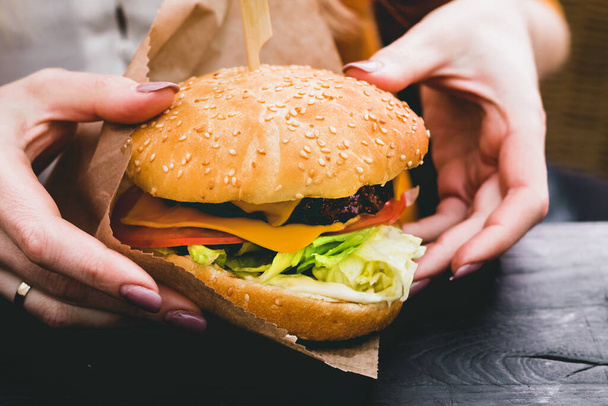 Weibliche Hände halten frische leckere Burger mit Pommes auf dem schwarzen Holztisch - Foto, Bild