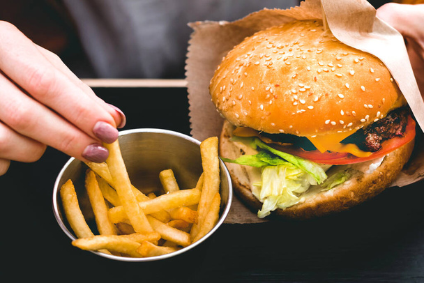 Vrouwelijke handen met verse heerlijke hamburger met frietjes op de zwarte houten tafel - Foto, afbeelding