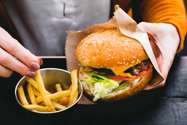Mains féminines tenant un délicieux burger frais avec des frites sur la table en bois noir - Photo, image