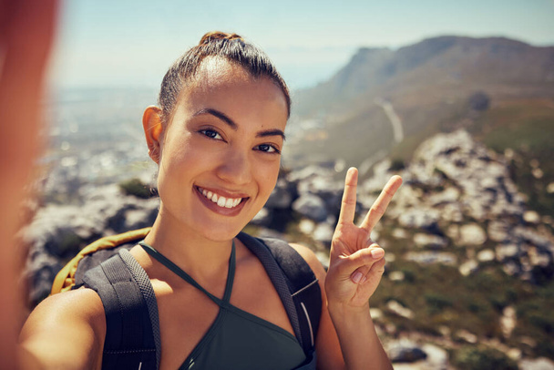 Selfie senderismo, mujer feliz y fitness en la naturaleza en una montaña para el ejercicio, viajes y aventura de trekking durante el verano. Cara, sonrisa y señal de paz turista femenina explorando al aire libre con una mochila. - Foto, Imagen