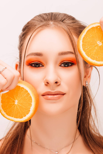 Das Mädchen hält zwei Hälften saftiger Orangen in der Nähe ihres Gesichts. Porträt eines jungen schönen Mädchens mit hellem Make-up und Orangen - Foto, Bild