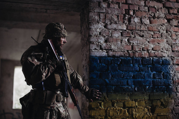 Ukrainan armeijan mies, jolla on ase käsissään, seisoo Ukrainan lipun alla. Ukrainan lippu on maalattu tiiliseinään. - Valokuva, kuva