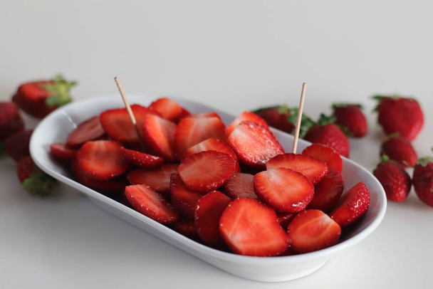 Μισό κομμένο φρέσκο φράουλες μέσα σε ένα λευκό μπολ, πυροβόλησε σε λευκό φόντο. Τα πιο διαδεδομένα μούρα στον κόσμο, που καλλιεργούνται εμπορικά παγκοσμίως ως γλυκά, αρωματικά και ζουμερά φρούτα. - Φωτογραφία, εικόνα