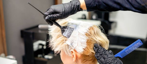 Раскрашивание седых волос краской для волос и расческой руками парикмахера для молодой белокурой белокурой женщины в парикмахерской, крупным планом - Фото, изображение