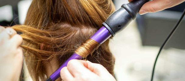 The hairstylist makes curls hairstyle of long brown hair with the curling iron in hairdresser salon, close up - Φωτογραφία, εικόνα