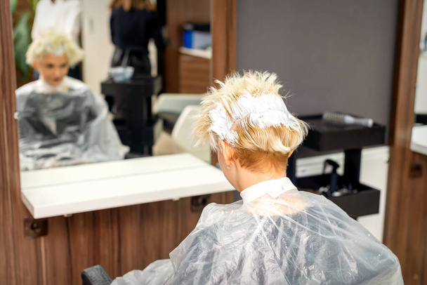 Coloriage cheveux blancs avec teinture capillaire de la jeune femme blonde caucasienne assise dans un salon de coiffure, gros plan - Photo, image