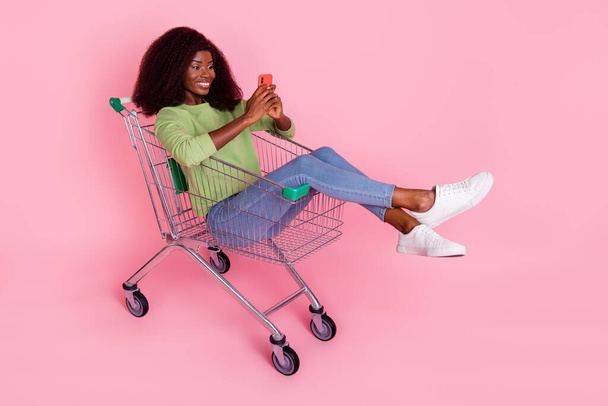Full length photo of pretty cute lady wear green sweater sitting shopping tray chatting gadget isolated pink color background. - Foto, Imagem