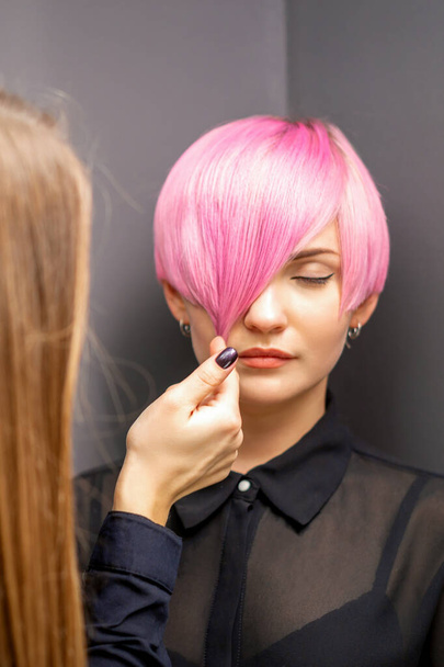 Hairdresser with hands is checking out and fixing the short pink hairstyle of the young white woman in a hair salon - Foto, Imagen
