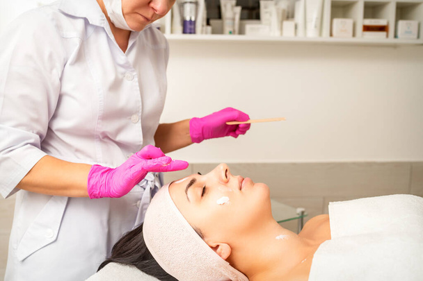 Cosmetologist with gloved hands applies a moisturizing mask with peeling cream on the female face. Facial cosmetology treatment. Procedures for facial care - Photo, Image