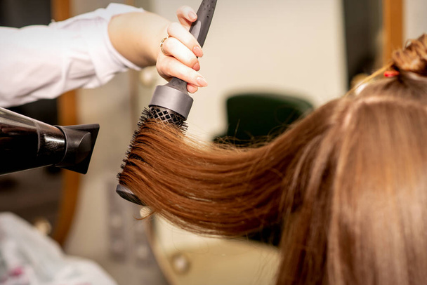 Ένα κομμωτήριο στεγνώνει μακριά καστανά μαλλιά με στεγνωτήρα μαλλιών και στρογγυλό πινέλο σε ένα σαλόνι ομορφιάς - Φωτογραφία, εικόνα