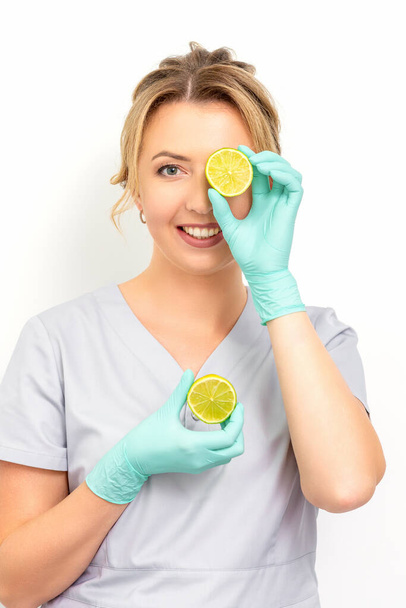 Portrait of young caucasian smiling female beautician covering eye with a lime slice wearing gloves over a white background - Photo, Image