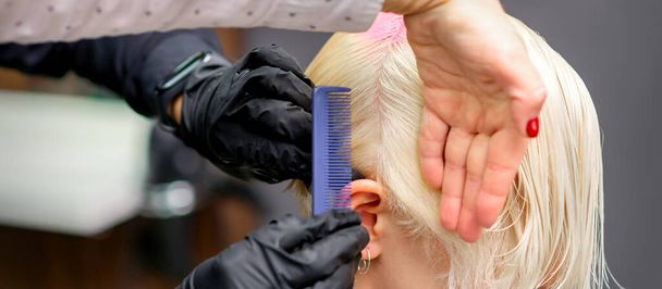 Χτένισμα μαλλιών με χτένα κατά τη βαφή άσπρα μαλλιά μιας νεαρής ξανθιάς γυναίκας στο κομμωτήριο - Φωτογραφία, εικόνα