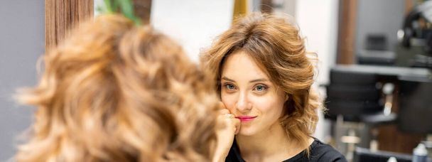 Die schöne junge kaukasische rothaarige Frau mit einer neuen kurzen welligen Frisur schaut in den Spiegel und überprüft Frisur und Make-up in einem Friseursalon - Foto, Bild