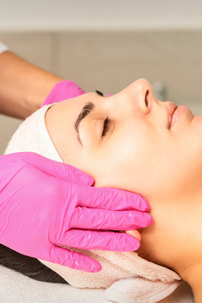 Kosmetologi käsineillä kädet soveltaa kosteuttava naamio kuorinta kerma naisen kasvot. Kasvojen kosmetiikkahoito. Kasvojen hoitoon liittyvät menettelyt - Valokuva, kuva