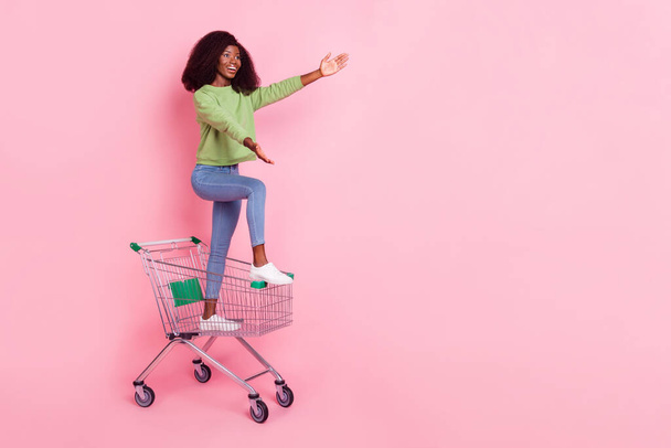 Full length photo of pretty sweet lady wear green sweater riding shopping tray arms empty space isolated pink color background. - 写真・画像