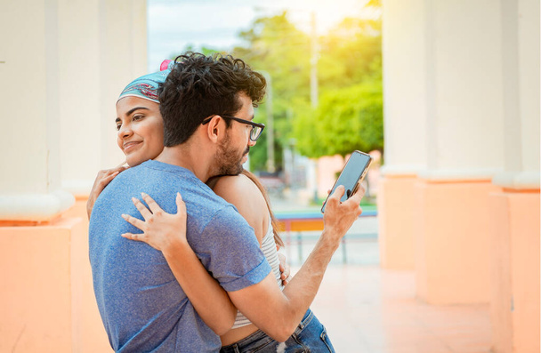 Ο άπιστος φίλος αγκαλιάζει την κοπέλα του και κοιτάει το κινητό. Έννοια της απιστίας του ανθρώπου χρησιμοποιώντας το κινητό τηλέφωνο. Απιστος ανθρωπος κοιταει το κινητο ενω αγκαλιαζει την κοπελα του εξω - Φωτογραφία, εικόνα