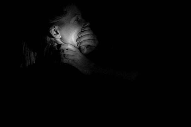 Το χέρι ενός άντρα κλείνει το στόμα του και στραγγαλίζει ένα κορίτσι στο σκοτάδι, βία και φόβο, βία εναντίον μιας γυναίκας, ενός δολοφόνου. - Φωτογραφία, εικόνα