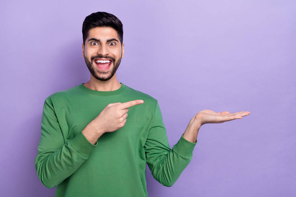 Photo of positive optimistic cheerful guy wear green pullover indicating empty space astonished staring isolated on violet color background. - Фото, изображение