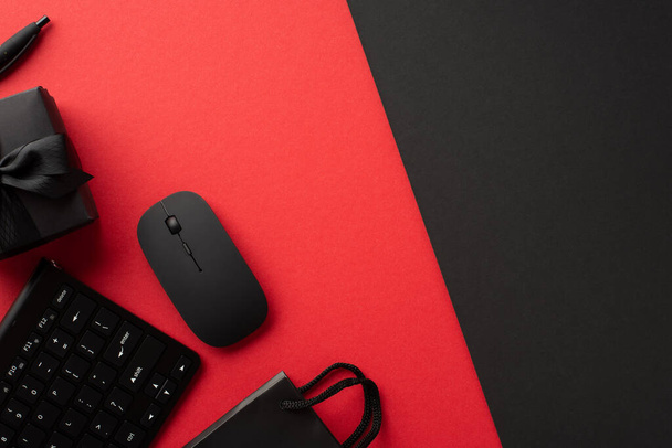 Koncept kybernetického pondělí. Horní pohled fotografie klávesnice počítač myš černá dárková krabice s mašlí luk pero a papírový sáček na bicolor červené a černé pozadí s prázdným prostorem - Fotografie, Obrázek