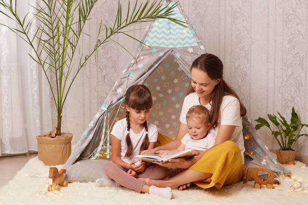 Horizontal shot of happy mother wearing white shirt and yellow trousers sitting on floor with her kids and reading book, being interested in interesting plot of fairy tale. - 写真・画像