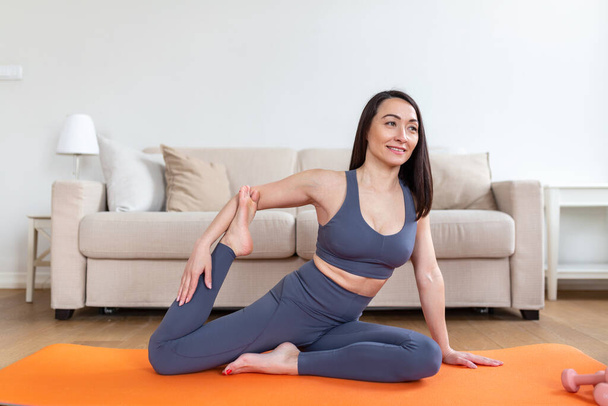 Joven mujer sonriente atractiva practicando yoga, sentada en pose de sirena, haciendo ejercicio, usando ropa deportiva, sesión de meditación, interior de cuerpo entero, interior del hogar - Foto, Imagen