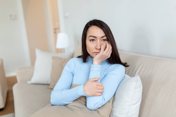 Frustrated Asian lady sitting on sofa, cuddling pillow, looking away at window. Lost in thoughts unhappy stressed woman regretting of wrong decision, spending time alone in living room. - Valokuva, kuva