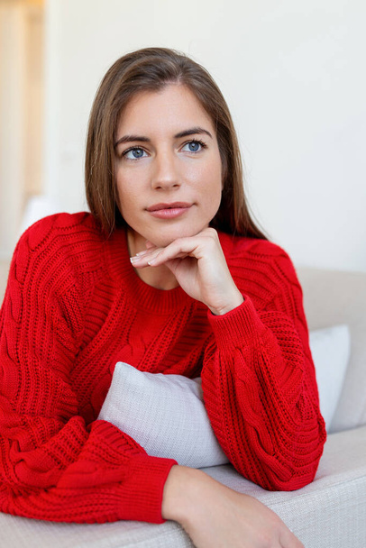 Happy young woman sitting on sofa at home and looking at camera. Portrait of comfortable woman in winter clothes relaxing on armchair. Portrait of beautiful woman smiling and relaxing during autumn. - Фото, изображение