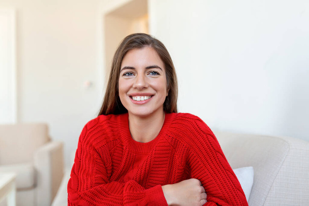 Happy young woman sitting on sofa at home and looking at camera. Portrait of comfortable woman in winter clothes relaxing on armchair. Portrait of beautiful woman smiling and relaxing during autumn. - Photo, Image