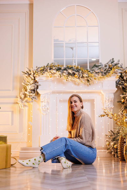 Una joven mujer feliz está sentada con un regalo de Año Nuevo. Árbol de Navidad y chimenea con guirnaldas festivas. El humor de Año Nuevo - Foto, Imagen