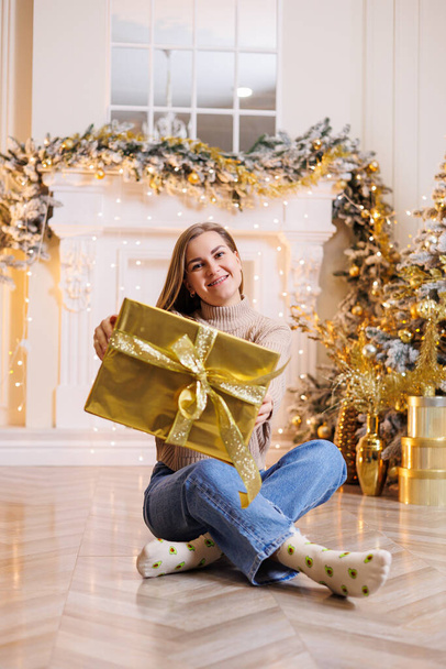 Молода щаслива жінка сидить з новорічним подарунком. Ялинка і камін зі святковими гірляндами. Новорічний настрій
 - Фото, зображення
