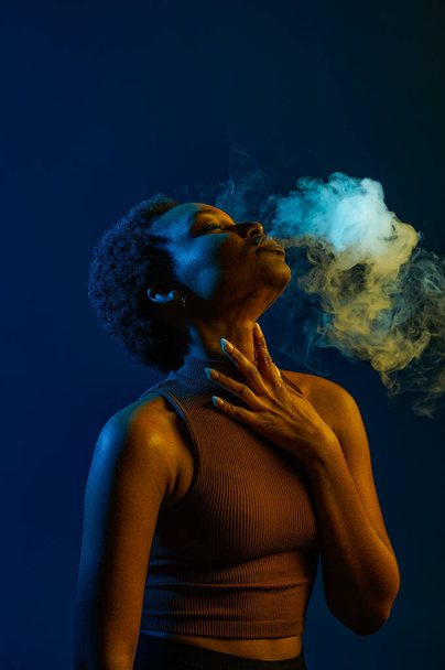 Passion woman blowing smoke curls from mouth over dark studio background in neon light - Foto, Bild