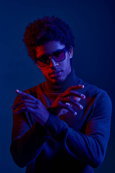 Junger selbstbewusster Mann mit Sonnenbrille, cooler Typ, der vor dunklem Hintergrund für ein Kamerastudio posiert - Foto, Bild