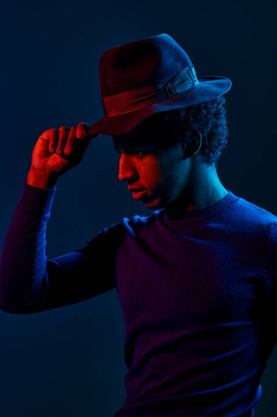 Młody człowiek w kapeluszu ze stylowym widokiem na ciemne tło studia pod oświetleniem neonowym - Zdjęcie, obraz