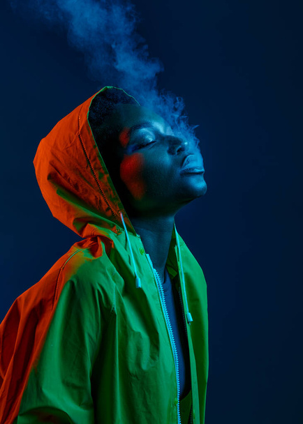 Hipster woman in raincoat blowing smoke from mouth over dark studio background under neon light illumination - Fotó, kép