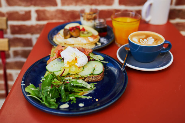 Delicious healthy brunch with avocado and salmon toast, boiled egg, pancakes with fruits and maple syrup and flat white coffee in cafe of Paris, France - Photo, Image