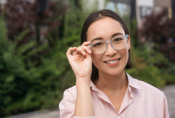 Portrait of smiling asian woman corrects stylish eyeglasses looking at camera standing outdoors  - Fotografie, Obrázek