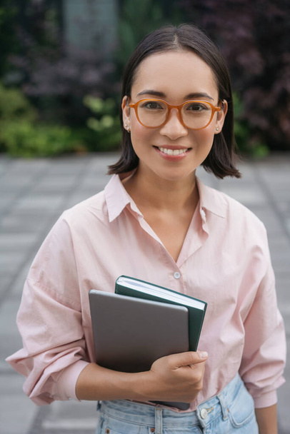 Retrato de estudiante asiática sonriente inteligente con gafas elegantes sosteniendo libro, portátil de pie en el campus universitario. Concepto educativo  - Foto, imagen
