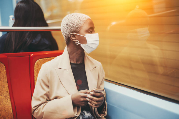 Profile view of an elegant African lady in a facial anti-virus protective mask sitting in a car of a metro train with a smartphone in her hands and looking outside the window at the motion - Foto, Imagem