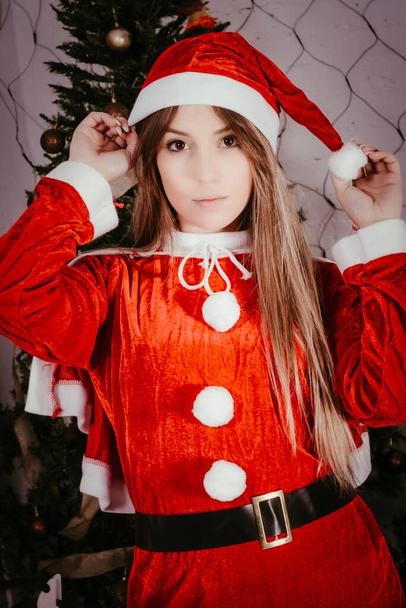 Nuori nainen pukeutunut punaiseen Mama Noelin asuun, jouluna joulukuusen edessä hattua pitelemässä. Joulukuuset. - Valokuva, kuva