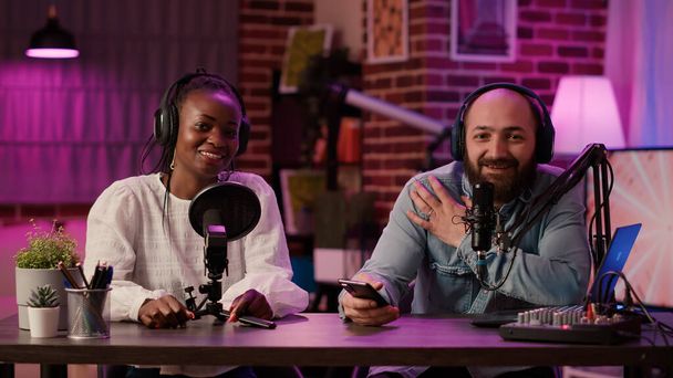 Diverse team of online radio hosts recording podcast review for smartphone streaming audio with digital mixer. Content creator and african american influencer broadcasting live product presentation. - Photo, Image