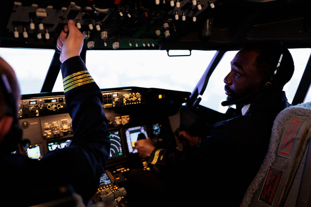 Pilotlardan oluşan çok ırklı bir ekip, pilot kabininde kontrol panelini kullanarak uçak uçuruyor, güç düğmesine basıyor ve kalkış için kontrol paneli düğmelerine basıyor. Kulübede uçuş ekibiyle birlikte uluslararası havayolu hizmeti. - Fotoğraf, Görsel