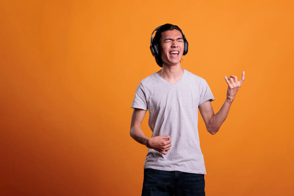 Young asian man in wireless, headphones playing virtual guitar, rockstar lifestyle. Teenage guitarist practicing, using imaginable musical instrument, fun pastime, leisure activity - 写真・画像