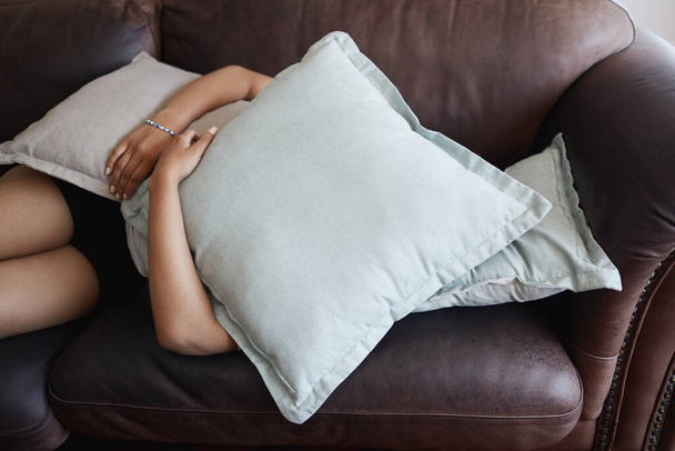 Tired, depressed girl sleeping under pillow on sofa in the living room. Woman hide feeling sad or anxiety and problem with depression, mental health and fatigue or fear on couch in the lounge. - Photo, Image