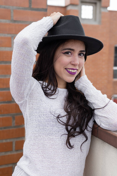 sosteniendo su sombrero una mujer latina con el pelo ondulado largo, accesorio de moda, belleza y maquillaje natural en el día, detalles de la cara - Foto, imagen