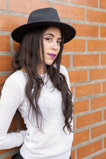 foto estúdio com o rosto de uma mulher latina com cabelo preto longo, usa uma maquiagem e chapéu, no fundo parede de tijolo, beleza e acessório à moda e juventude - Foto, Imagem