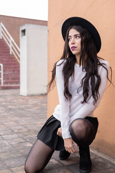 beautiful latin young woman with long hair posing squatting wearing a hat, sweater, skirt and tights, fashion and beauty with makeup, lifestyle - Foto, imagen