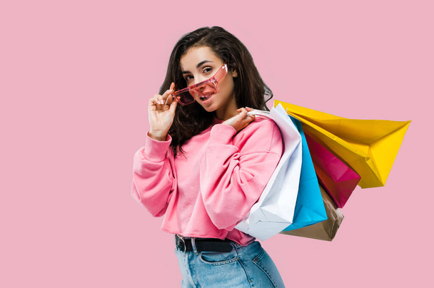 Photo of beautiful caucasian stylish brunette young woman, wearing casual clothes and pink sunglasses, holding shopping bags, stand on isolated pink background, looks at camera, smile.Shopping concept - Valokuva, kuva