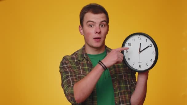 It is your time. Portrait of teen young man in shirt showing time on clock watch, ok, thumb up, approve, pointing finger at camera. Adult boy indoors studio shot isolated alone on yellow background - Footage, Video